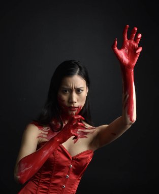 Kırmızı korse giyen güzel Asyalı bir kadının yakın plan portresi, kanlı boyayla kaplanmış, Gotik korku tarzında, yaratıcı el hareketleriyle koyu siyah stüdyo arka planına izole edilmiş..