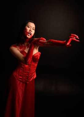 Kırmızı korse giyen güzel Asyalı bir kadının yakın plan portresi, kanlı boyayla kaplanmış, Gotik korku tarzında, yaratıcı el hareketleriyle koyu siyah stüdyo arka planına izole edilmiş..