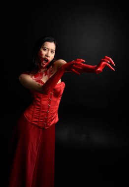 Kırmızı korse giyen güzel Asyalı bir kadının yakın plan portresi, kanlı boyayla kaplanmış, Gotik korku tarzında, yaratıcı el hareketleriyle koyu siyah stüdyo arka planına izole edilmiş..