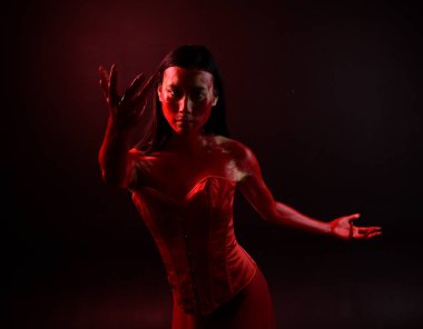 Kırmızı korse giyen güzel Asyalı bir kadının yakın plan portresi, kanlı boyayla kaplanmış, Gotik korku tarzında, yaratıcı el hareketleriyle koyu siyah stüdyo arka planına izole edilmiş..