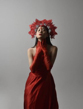 Close up portrait of beautiful young asian woman wearing red corset, long opera gloves and ornate crown headdress. Gestural hand poses isolated on light studio background.