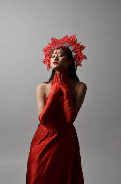 Close up portrait of beautiful young asian woman wearing red corset, long opera gloves and ornate crown headdress. Gestural hand poses isolated on light studio background.