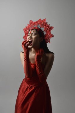Close up portrait of beautiful young asian woman wearing red corset, long opera gloves and ornate crown headdress. Gestural hand poses isolated on light studio background.