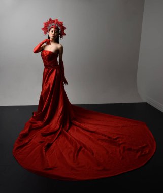 Full length  portrait of beautiful young asian woman wearing red corset, long opera gloves and ornate crown headdress. Gestural hand poses isolated on light studio background.