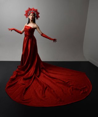 Full length  portrait of beautiful young asian woman wearing red corset, long opera gloves and ornate crown headdress. Gestural hand poses isolated on light studio background.