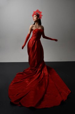 Full length  portrait of beautiful young asian woman wearing red corset, long opera gloves and ornate crown headdress. Gestural hand poses isolated on light studio background.