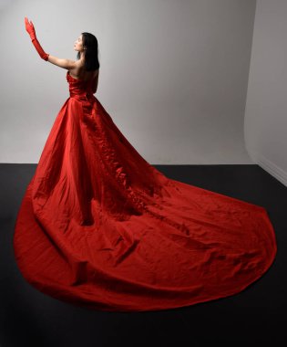 Full length  portrait of beautiful young asian woman wearing red corset, long opera gloves and ornate crown headdress. Gestural hand poses isolated on light studio background.