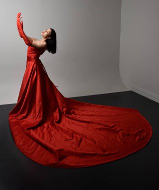 Full length  portrait of beautiful young asian woman wearing red corset, long opera gloves and ornate crown headdress. Gestural hand poses isolated on light studio background.