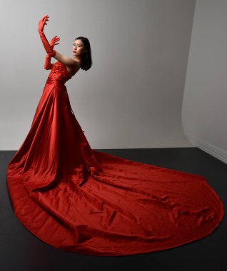 Full length  portrait of beautiful young asian woman wearing red corset, long opera gloves and ornate crown headdress. Gestural hand poses isolated on light studio background.