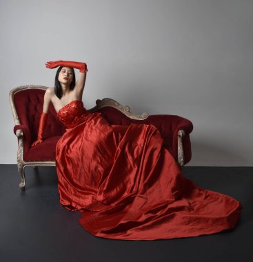 Full length  portrait of beautiful young asian woman wearing red corset, long opera gloves and ornate crown headdress. Sitting pose with gestural hands  isolated on light studio background.