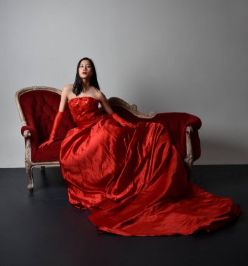 Full length  portrait of beautiful young asian woman wearing red corset, long opera gloves and ornate crown headdress. Sitting pose with gestural hands  isolated on light studio background.