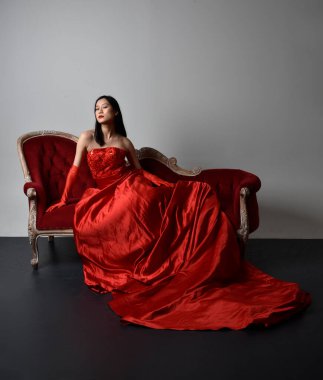 Full length  portrait of beautiful young asian woman wearing red corset, long opera gloves and ornate crown headdress. Sitting pose with gestural hands  isolated on light studio background.
