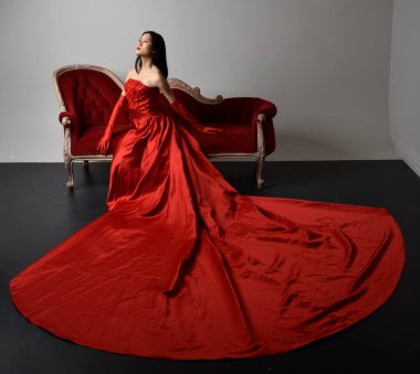 Full length  portrait of beautiful young asian woman wearing red corset, long opera gloves and ornate crown headdress. Sitting pose with gestural hands  isolated on light studio background.