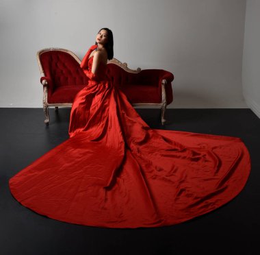 Full length  portrait of beautiful young asian woman wearing red corset, long opera gloves and ornate crown headdress. Sitting pose with gestural hands  isolated on light studio background.