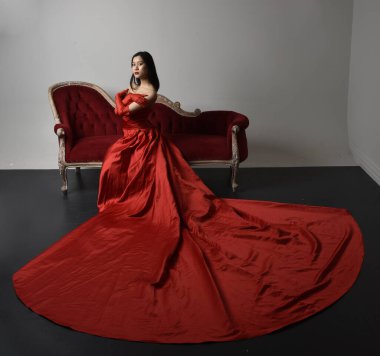 Full length  portrait of beautiful young asian woman wearing red corset, long opera gloves and ornate crown headdress. Sitting pose with gestural hands  isolated on light studio background.