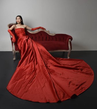 Full length  portrait of beautiful young asian woman wearing red corset, long opera gloves and ornate crown headdress. Sitting pose with gestural hands  isolated on light studio background.