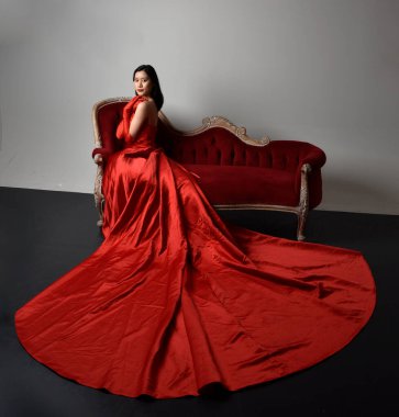 Full length  portrait of beautiful young asian woman wearing red corset, long opera gloves and ornate crown headdress. Sitting pose with gestural hands  isolated on light studio background.