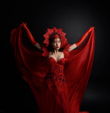 portrait of beautiful young asian woman wearing red corset, long opera gloves and ornate crown headdress. Sitting pose with gestural hands  isolated on light studio background.