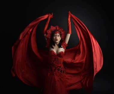 portrait of beautiful young asian woman wearing red corset, long opera gloves and ornate crown headdress. Sitting pose with gestural hands  isolated on light studio background.