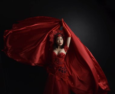 portrait of beautiful young asian woman wearing red corset, long opera gloves and ornate crown headdress. Sitting pose with gestural hands  isolated on light studio background.