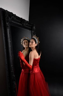 Kırmızı korse, uzun opera eldivenleri ve süslü taç başlık giyen Asyalı güzel bir kadının tam boy portresi. Stüdyonun arka planında uzun bir aynaya karşı zarif bir poz..