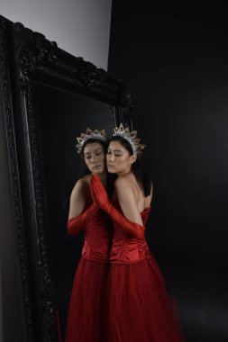 Kırmızı korse, uzun opera eldivenleri ve süslü taç başlık giyen Asyalı güzel bir kadının tam boy portresi. Stüdyonun arka planında uzun bir aynaya karşı zarif bir poz..