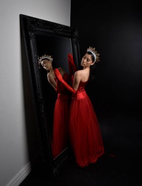 Kırmızı korse, uzun opera eldivenleri ve süslü taç başlık giyen Asyalı güzel bir kadının tam boy portresi. Stüdyonun arka planında uzun bir aynaya karşı zarif bir poz..