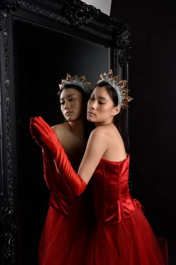 Kırmızı korse, uzun opera eldivenleri ve süslü taç başlık giyen Asyalı güzel bir kadının tam boy portresi. Stüdyonun arka planında uzun bir aynaya karşı zarif bir poz..