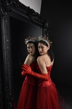 Kırmızı korse, uzun opera eldivenleri ve süslü taç başlık giyen Asyalı güzel bir kadının tam boy portresi. Stüdyonun arka planında uzun bir aynaya karşı zarif bir poz..