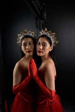 Kırmızı korse, uzun opera eldivenleri ve süslü taç başlık giyen Asyalı güzel bir kadının tam boy portresi. Stüdyonun arka planında uzun bir aynaya karşı zarif bir poz..