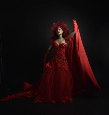 Kırmızı korse, uzun opera eldivenleri ve süslü gotik kraliçe tacı giyen güzel Asyalı bir kadının dramatik portresi. Karanlık bir stüdyo arka planında zarif bir poz..
