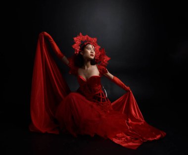 Kırmızı korse, uzun opera eldivenleri ve süslü gotik kraliçe tacı giyen güzel Asyalı bir kadının dramatik portresi. Karanlık bir stüdyo arka planında zarif bir poz..