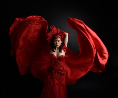 Kırmızı korse, uzun opera eldivenleri ve süslü gotik kraliçe tacı giyen güzel Asyalı bir kadının dramatik portresi. Karanlık bir stüdyo arka planında zarif bir poz..