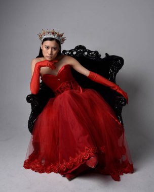 Kırmızı korse, uzun opera eldivenleri ve süslü gotik kraliçe tacı giyen Asyalı güzel bir kadının tam boy portresi. Sandalyede otururken zarif pozlar, stüdyo arka planında izole edilmiş..