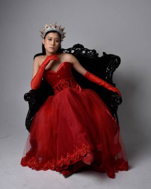 Kırmızı korse, uzun opera eldivenleri ve süslü gotik kraliçe tacı giyen Asyalı güzel bir kadının tam boy portresi. Sandalyede otururken zarif pozlar, stüdyo arka planında izole edilmiş..