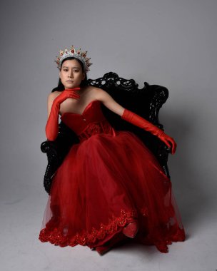 Kırmızı korse, uzun opera eldivenleri ve süslü gotik kraliçe tacı giyen Asyalı güzel bir kadının tam boy portresi. Sandalyede otururken zarif pozlar, stüdyo arka planında izole edilmiş..