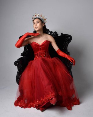 Kırmızı korse, uzun opera eldivenleri ve süslü gotik kraliçe tacı giyen Asyalı güzel bir kadının tam boy portresi. Sandalyede otururken zarif pozlar, stüdyo arka planında izole edilmiş..