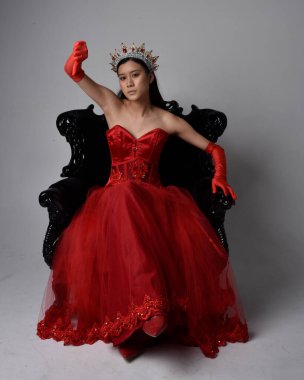 Kırmızı korse, uzun opera eldivenleri ve süslü gotik kraliçe tacı giyen Asyalı güzel bir kadının tam boy portresi. Sandalyede otururken zarif pozlar, stüdyo arka planında izole edilmiş..