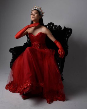 Kırmızı korse, uzun opera eldivenleri ve süslü gotik kraliçe tacı giyen Asyalı güzel bir kadının tam boy portresi. Sandalyede otururken zarif pozlar, stüdyo arka planında izole edilmiş..