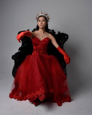 Kırmızı korse, uzun opera eldivenleri ve süslü gotik kraliçe tacı giyen Asyalı güzel bir kadının tam boy portresi. Sandalyede otururken zarif pozlar, stüdyo arka planında izole edilmiş..