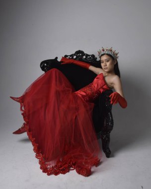 Kırmızı korse, uzun opera eldivenleri ve süslü gotik kraliçe tacı giyen Asyalı güzel bir kadının tam boy portresi. Sandalyede otururken zarif pozlar, stüdyo arka planında izole edilmiş..