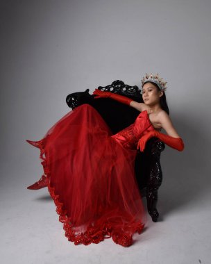 Kırmızı korse, uzun opera eldivenleri ve süslü gotik kraliçe tacı giyen Asyalı güzel bir kadının tam boy portresi. Sandalyede otururken zarif pozlar, stüdyo arka planında izole edilmiş..