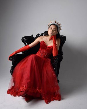 Kırmızı korse, uzun opera eldivenleri ve süslü gotik kraliçe tacı giyen Asyalı güzel bir kadının tam boy portresi. Sandalyede otururken zarif pozlar, stüdyo arka planında izole edilmiş..