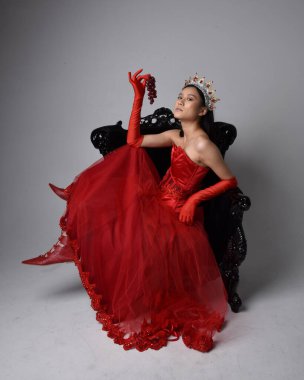 Kırmızı korse, uzun opera eldivenleri ve süslü gotik kraliçe tacı giyen Asyalı güzel bir kadının tam boy portresi. Sandalyede otururken zarif pozlar, stüdyo arka planında izole edilmiş..
