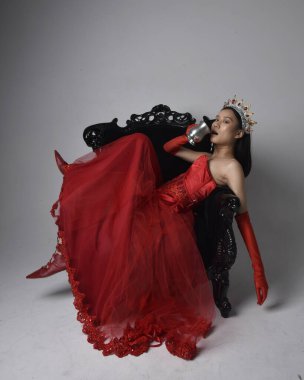 Kırmızı korse, uzun opera eldivenleri ve süslü gotik kraliçe tacı giyen Asyalı güzel bir kadının tam boy portresi. Sandalyede otururken zarif pozlar, stüdyo arka planında izole edilmiş..