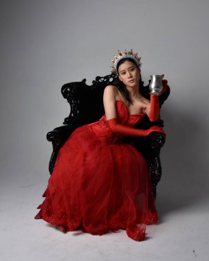 Kırmızı korse, uzun opera eldivenleri ve süslü gotik kraliçe tacı giyen Asyalı güzel bir kadının tam boy portresi. Sandalyede otururken zarif pozlar, stüdyo arka planında izole edilmiş..