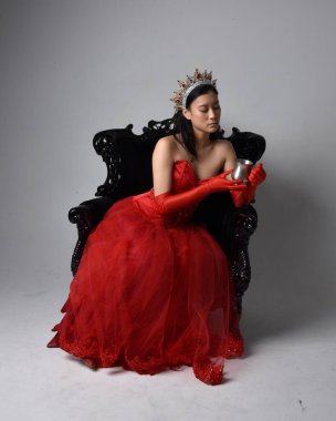 Kırmızı korse, uzun opera eldivenleri ve süslü gotik kraliçe tacı giyen Asyalı güzel bir kadının tam boy portresi. Sandalyede otururken zarif pozlar, stüdyo arka planında izole edilmiş..