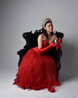 Kırmızı korse, uzun opera eldivenleri ve süslü gotik kraliçe tacı giyen Asyalı güzel bir kadının tam boy portresi. Sandalyede otururken zarif pozlar, stüdyo arka planında izole edilmiş..