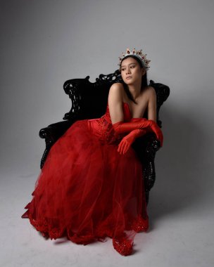 Kırmızı korse, uzun opera eldivenleri ve süslü gotik kraliçe tacı giyen Asyalı güzel bir kadının tam boy portresi. Sandalyede otururken zarif pozlar, stüdyo arka planında izole edilmiş..