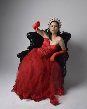 Kırmızı korse, uzun opera eldivenleri ve süslü gotik kraliçe tacı giyen Asyalı güzel bir kadının tam boy portresi. Sandalyede otururken zarif pozlar, stüdyo arka planında izole edilmiş..